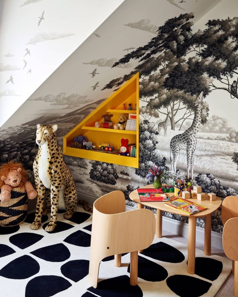 Cantinho com papel de parede de animais, com cores vibrantes, uma mesa e cadeira de madeira com alguns livros e brinquedos, onde pode ser o local de estudo para as crianças.